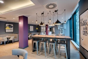 A modern dining area with a central island, bar stools, and a view of the city through large windows. - Premier Inn Berlin Kurfürstendamm (Berlin)