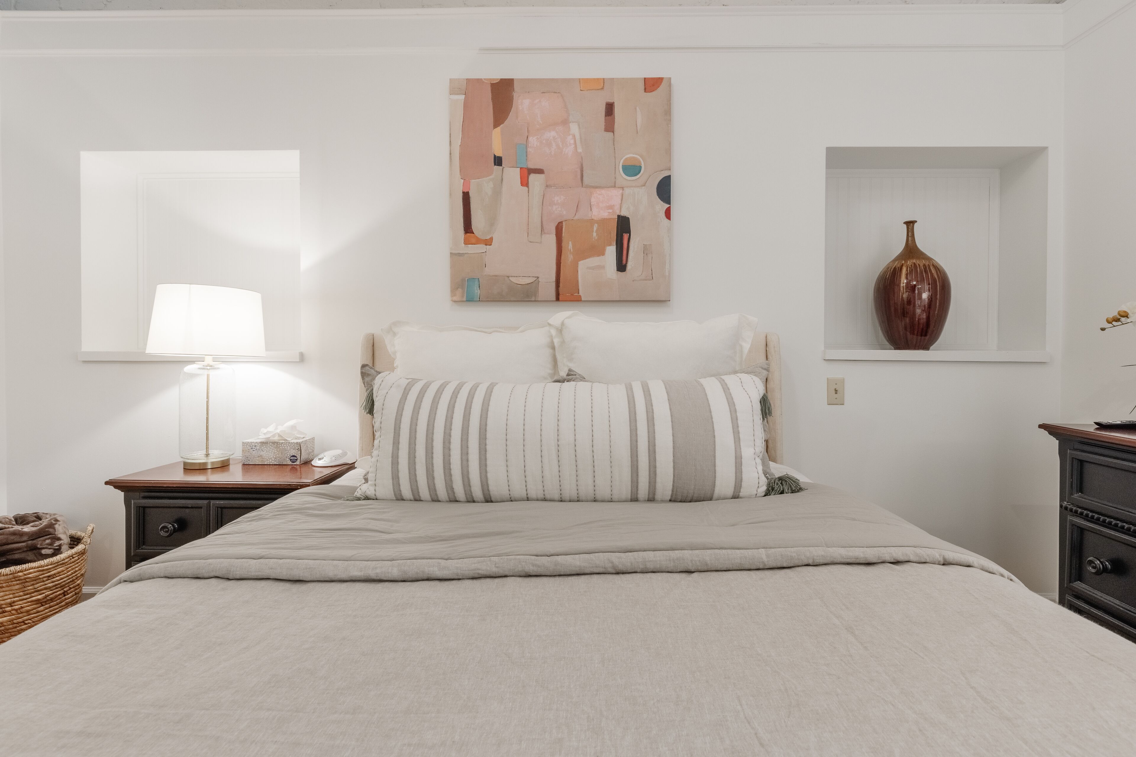 Een slaapkamer met een bed, een nachtkastje met een lamp, een wanddecoratie in abstractie en een vaas op een plank.