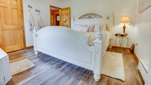 Une chambre avec un lit blanc, un plancher en bois, un tapis blanc, une table de chevet avec une lampe et une porte sur laquelle sont accrochées des serviettes.