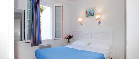 A bedroom with a bed, a window with curtains, a nightstand with a vase of flowers, and wall-mounted lights.