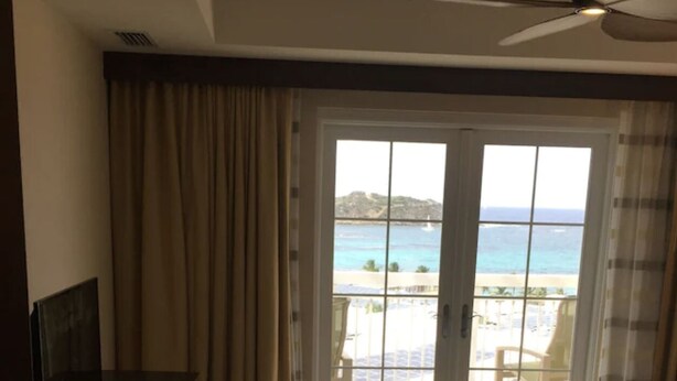 A hotel room with a bed, a TV, a ceiling fan, and a view of the beach through the window. - Amazing 3 bedroom condo (Philipsburg)