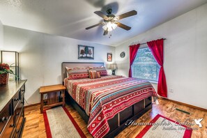 Ein Schlafzimmer mit einem hölzernen Bett, einem Deckenventilator, einem Fenster mit roten Vorhängen und einem Nachttisch mit einer Lampe.