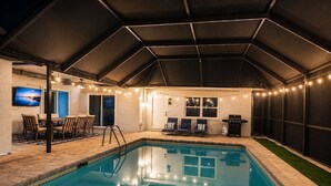 Una piscina con un porche cubierto, una parrilla y luces colgantes.