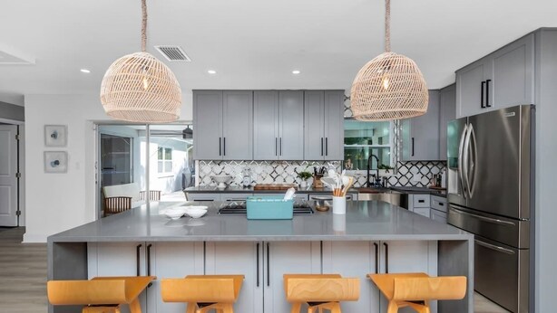 Una cocina moderna con gabinetes grises, una isla central con taburetes de bar, electrodomésticos de acero inoxidable y luminarias colgantes tejidas.