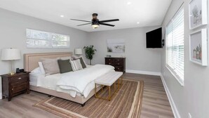 Un dormitorio con cama, mesitas de noche, ventilador de techo, televisor y obras de arte en las paredes.