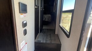 A narrow hallway with a wooden staircase leading to a bed, a control panel, and a window with a view.