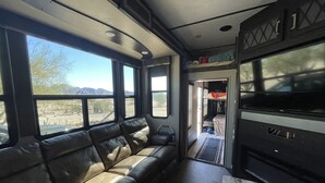 A compact RV interior with a leather sofa, a kitchenette, and a large window offering a view of the outdoors.