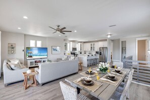 A spacious living area with a sofa, dining table set for four, a kitchen in the background, and a balcony with a railing.