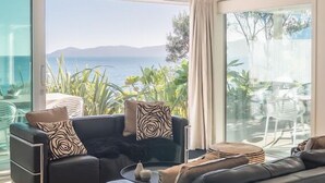 A modern living room with a large glass door leading to a balcony, a black sofa with patterned cushions, and a small round table with decorative items. - Seascapes Waterfront 2 (Paraparaumu)