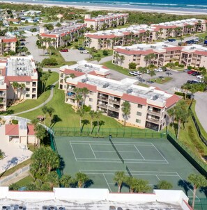 Vue aérienne d’un complexe résidentiel comprenant plusieurs immeubles d’appartements, un court de tennis et une plage en arrière-plan.