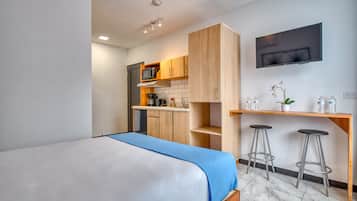 Quarto de hotel moderno com uma cama grande, uma cozinha compacta, uma televisão na parede e um pequeno bar com banquetas.