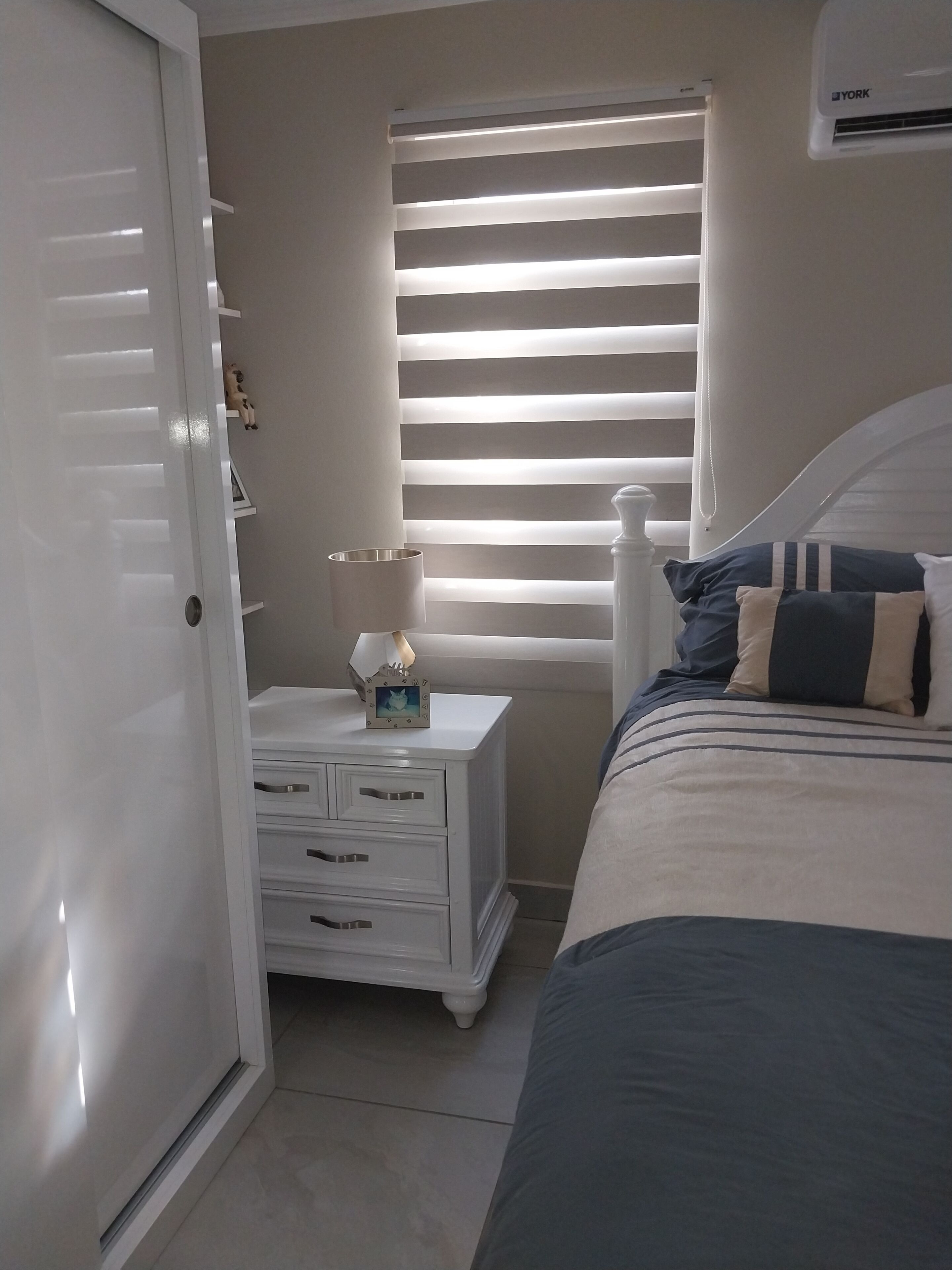 A bedroom with a bed, a nightstand with drawers, a lamp, a picture frame, and a window with blinds.