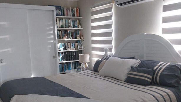 Un dormitorio con cama, estantería, aire acondicionado y ventana con persianas.