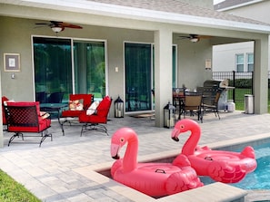 A pool area with two pink flamingo floats, red chairs, a table with chairs, and a grill.
