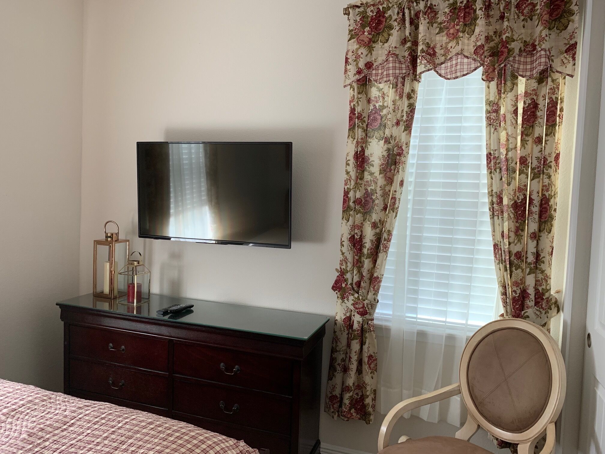 Un dormitorio con una televisión de pantalla plana montada en la pared, un aparador de madera con cajones, una cama con colcha a cuadros y una silla con asiento acolchado.