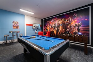 Une table de billard recouverte d’un drap bleu, entourée de tabourets et d’un canapé rouge. Une grande affiche de film est accrochée au mur derrière la table.