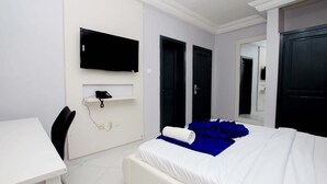 A hotel room with a bed, a desk, a chair, a TV mounted on the wall, and two doors. - Hotel Galilée (Cotonou)