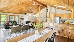 Un'ampia zona giorno con soffitto in legno, un tavolo da pranzo con sedie, una sala da conversazione e un angolo cottura con pensili in legno e un frigorifero in acciaio inossidabile.