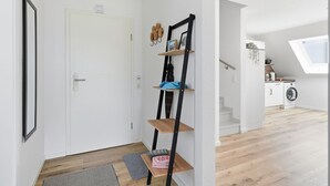 A modern interior with a white door, a black metal ladder shelf, a grey rug, and a view of a kitchen with a washing machine.