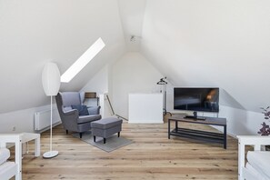 A modern loft bedroom with a sloped ceiling, a large TV, a grey sofa, and a wooden floor.