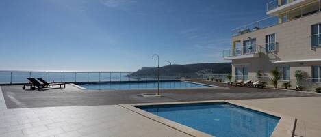 A rooftop pool area with two pools, lounge chairs, and a view of the sea.