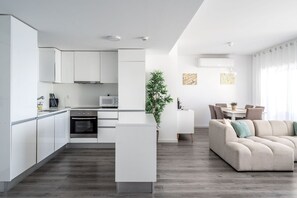 Eine moderne Küche mit weißen Schränken, ein Essbereich mit einem beigen Sofa und ein Wohnzimmer mit einem großen Fenster.