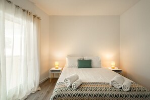 Ein Schlafzimmer mit einem Bett, zwei Nachttischlampen, einem Fenster mit Vorhängen und einer gemusterten Bettdecke.