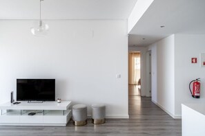Sala de estar moderna com televisão de tela plana, console branco, dois pufes cinzas e um extintor de incêndio à vista.