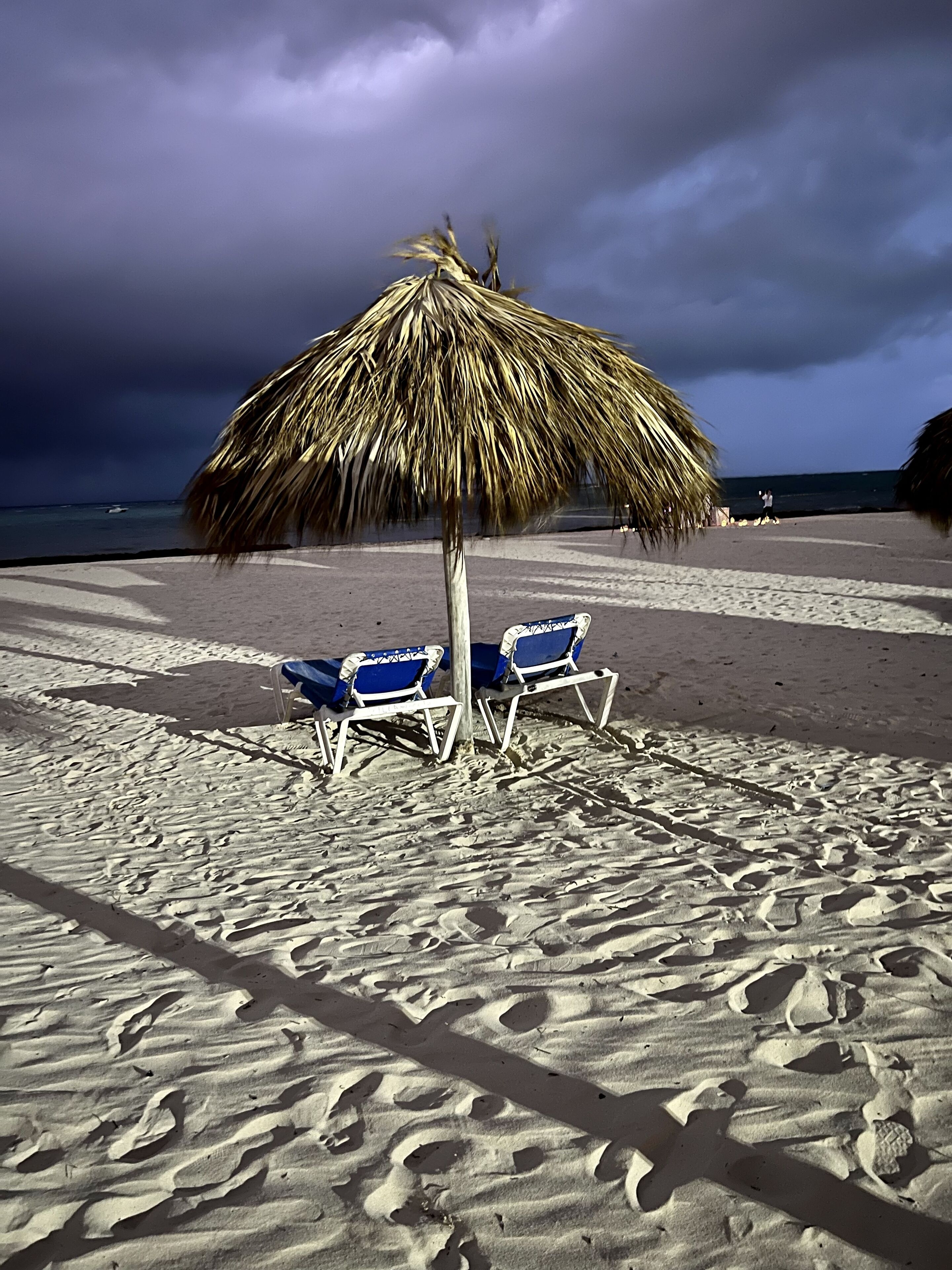 Une plage avec deux chaises longues bleues sous un parasol en chaume, une plage de sable et l’océan en arrière-plan.