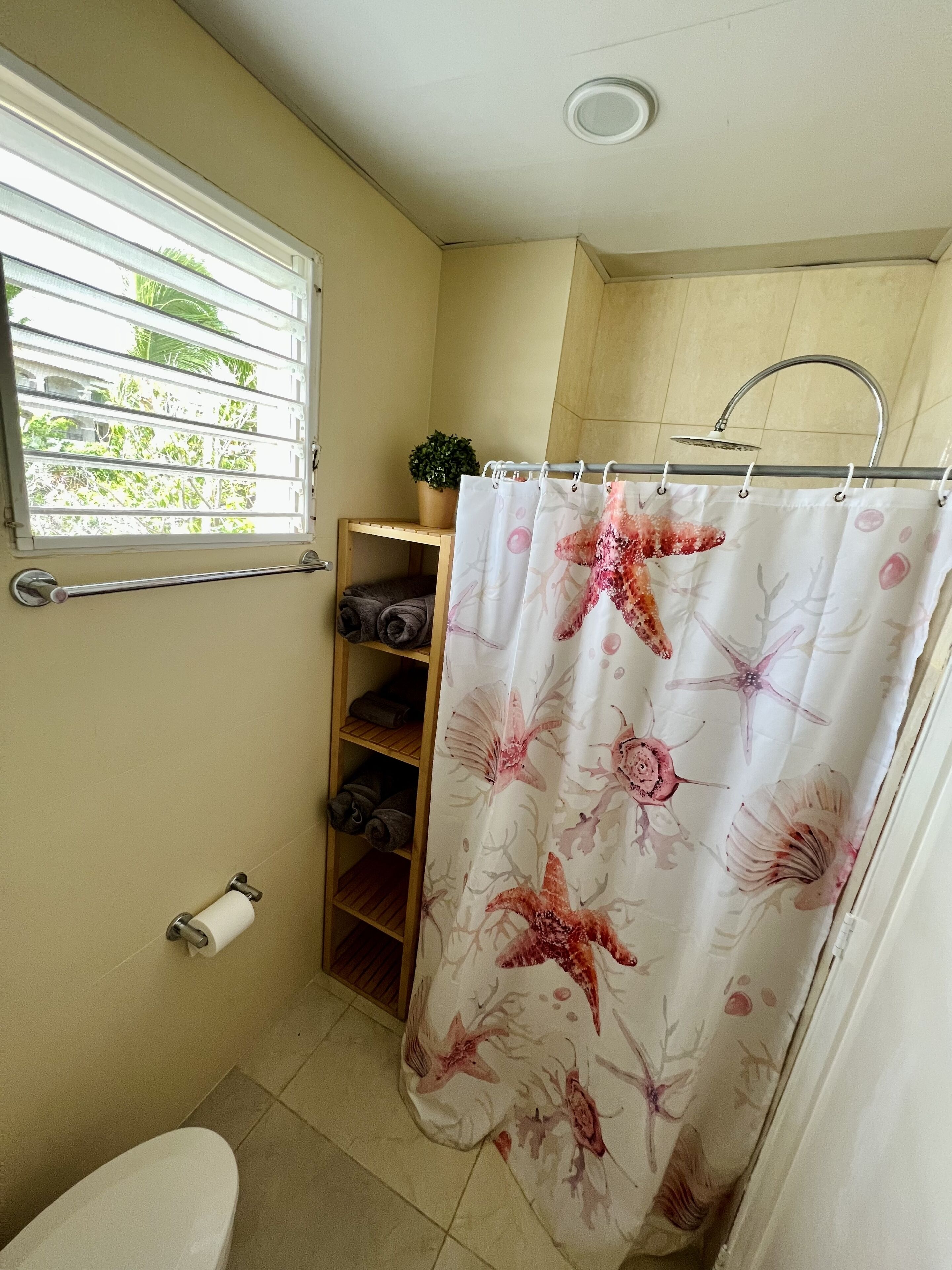 Un bagno con tenda da doccia a motivi floreali, un water, un porta asciugamani, uno scaffale con asciugamani piegati e una finestra con persiane.