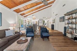 A spacious living area with a high ceiling, wooden beams, a sofa, two armchairs, a coffee table, and a bookshelf.