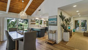 Een moderne keuken met een centraal eiland, een eethoek met een tafel en stoelen, en een woonruimte met een grote plant en kunstwerken.