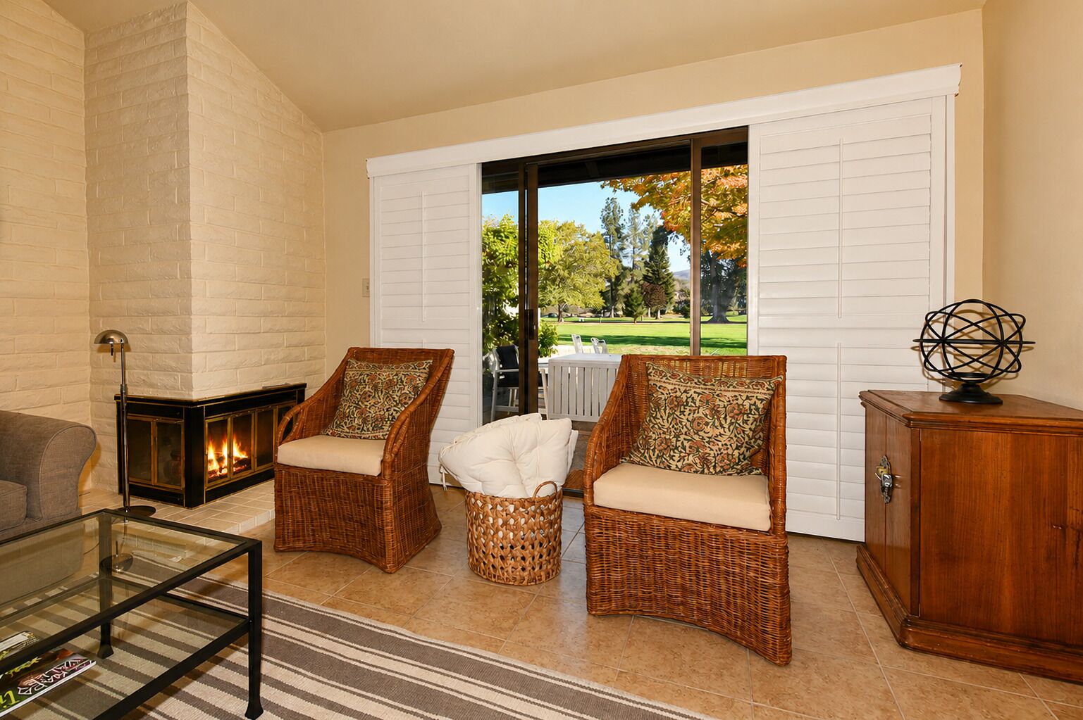 A living room with wicker chairs, a glass coffee table, a fireplace, and a view through a sliding glass door.