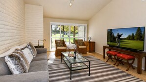 Ein Wohnzimmer mit Sofa, Sesseln, Couchtisch, Fernseher und einem großen Fenster mit Jalousien.