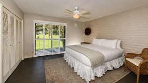 Ein Schlafzimmer mit einem Bett, einem Deckenventilator, einem Korbstuhl und Blick auf eine grüne Rasenfläche durch eine Schiebetür.