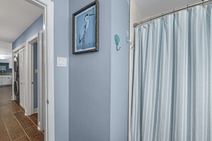 A hallway with a light blue wall, a framed painting of a bird, a shower curtain, and a door with a towel rack. - Gulf Village 109 | Direct Beach Access | Outdoor Pool | Ocean View | My Beach Getaways (Gulf Shores)