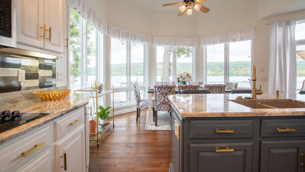 Cocina con una isla grande, piso de madera y vistas al exterior. Hay armarios con tiradores dorados, un ventilador de techo y un comedor con vistas.