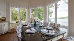 Un comedor con una mesa dispuesta para seis personas, amplias ventanas con vista a un lago y una cocina con muebles blancos.