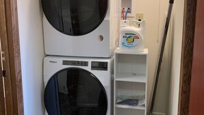 Une buanderie équipée de deux lave-linge Whirlpool superposés, d’un seau rouge et d’une étagère contenant des produits de nettoyage.