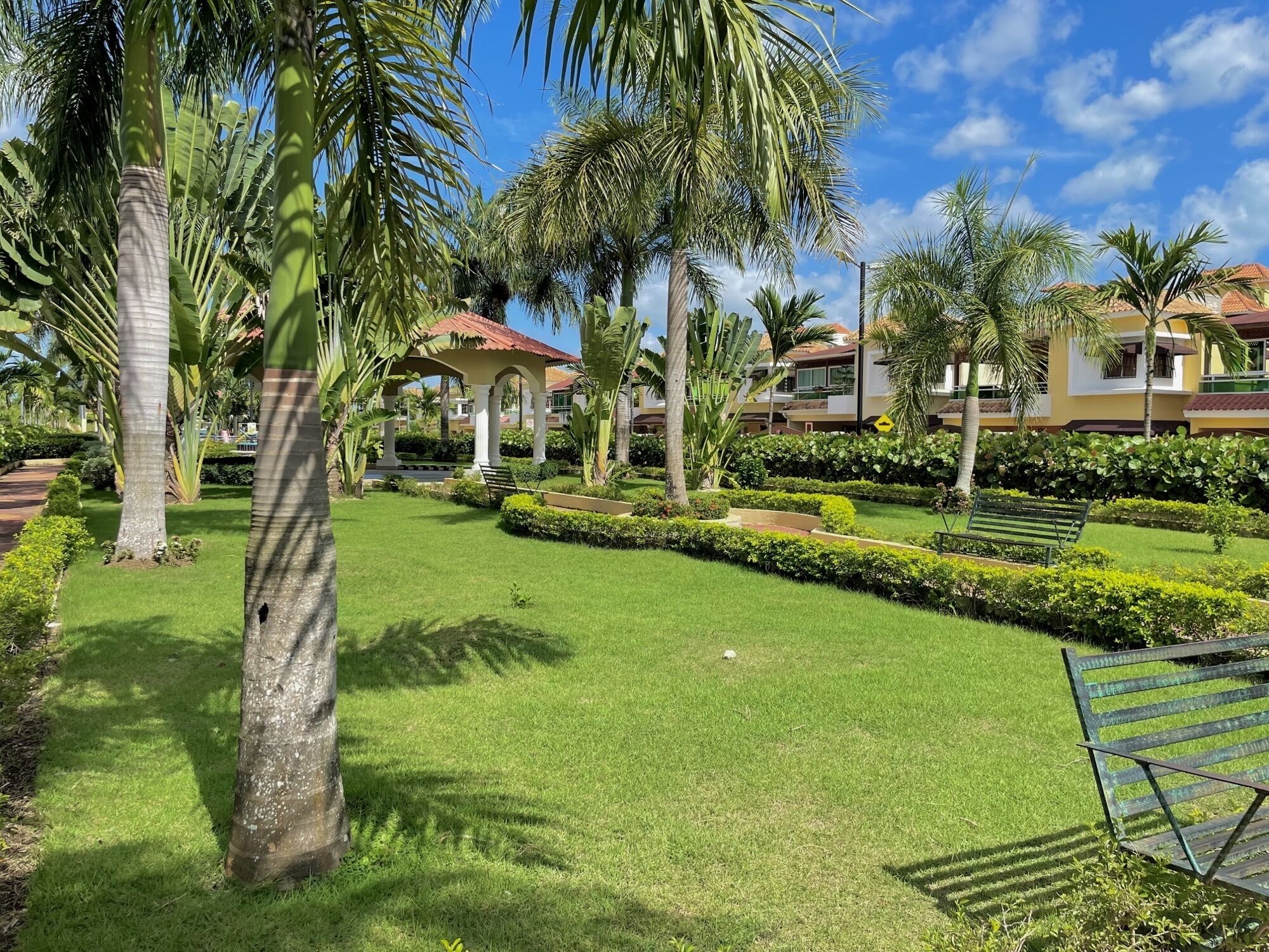 A well-maintained garden with palm trees, a gazebo, and neatly trimmed hedges.