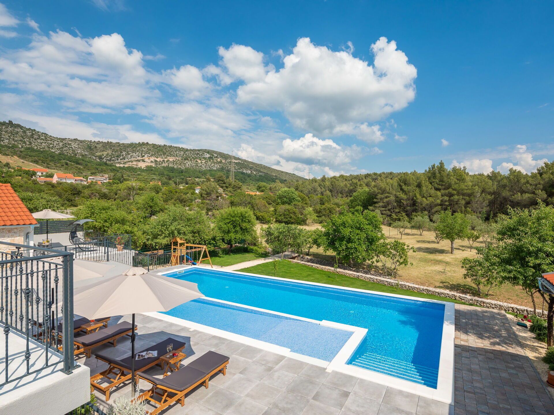 A swimming pool with lounge chairs and a wooden play structure, surrounded by a landscaped garden and a view of a hilly landscape.