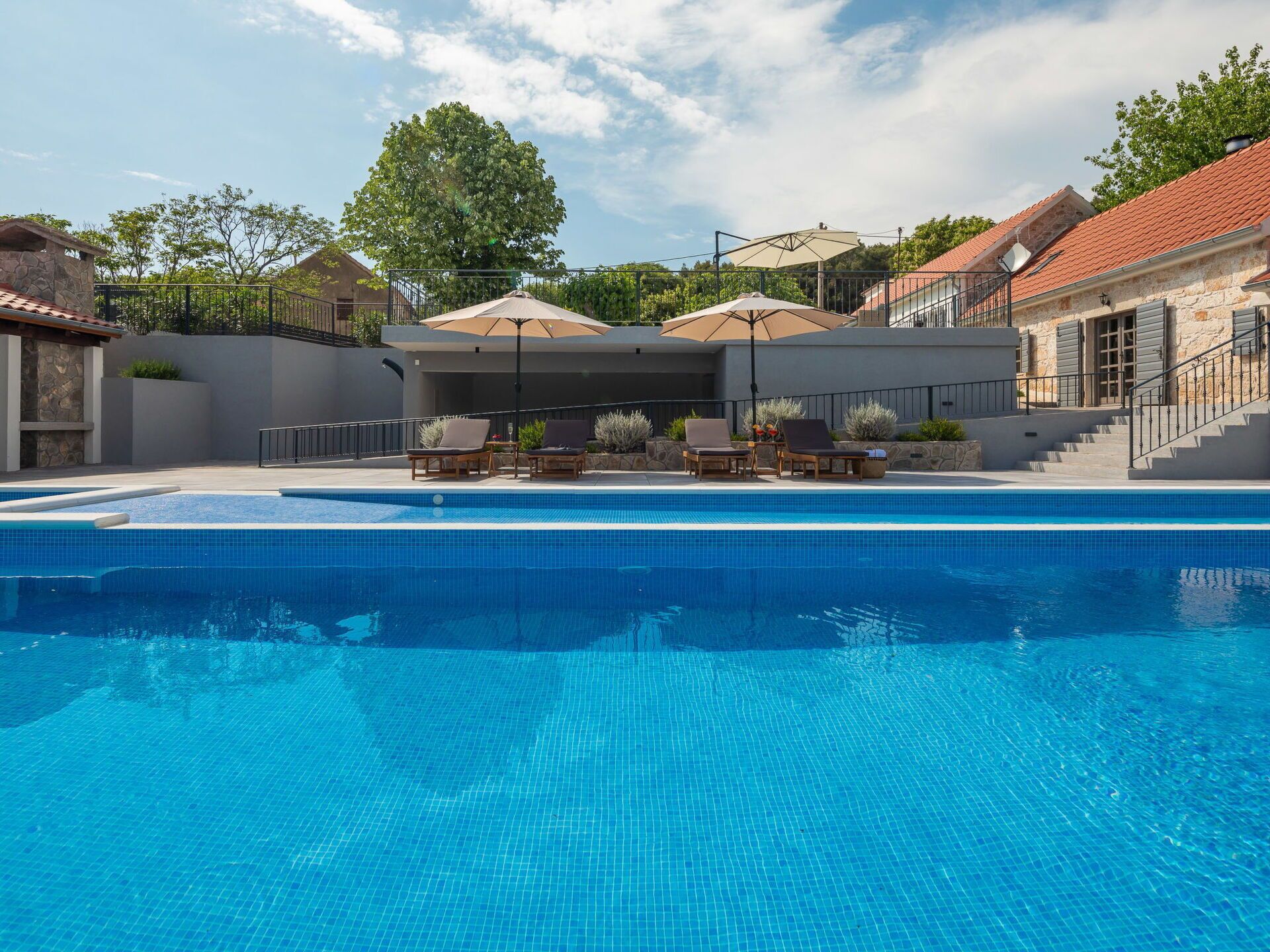 Une piscine entourée de bâtiments en pierre et d’un grand arbre, avec des chaises longues et des parasols à proximité.