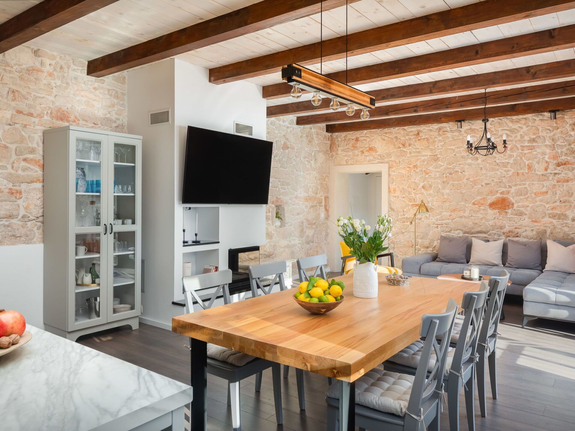 A modern living room with exposed wooden beams, a stone wall, a flat-screen TV, a dining table with chairs, and a sofa.