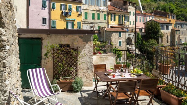 A terrace with a table set for dining, surrounded by stone walls and colorful buildings. - Nice apartment for 6 guests with terrace and pets allowed (Dolcedo)