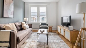 A modern living room with a sofa, coffee table, TV, and a view of the city through large windows. - Bright Horgen 2.5 Room w/ Storage, 3 min to Market, by Blueground (Zurich)