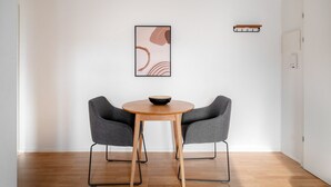 A minimalist room with a round wooden table, two grey upholstered chairs, a framed abstract artwork, and a small shelf with a book. - Bright Horgen 2.5 Room w/ Storage, 3 min to Market, by Blueground (Zurich)