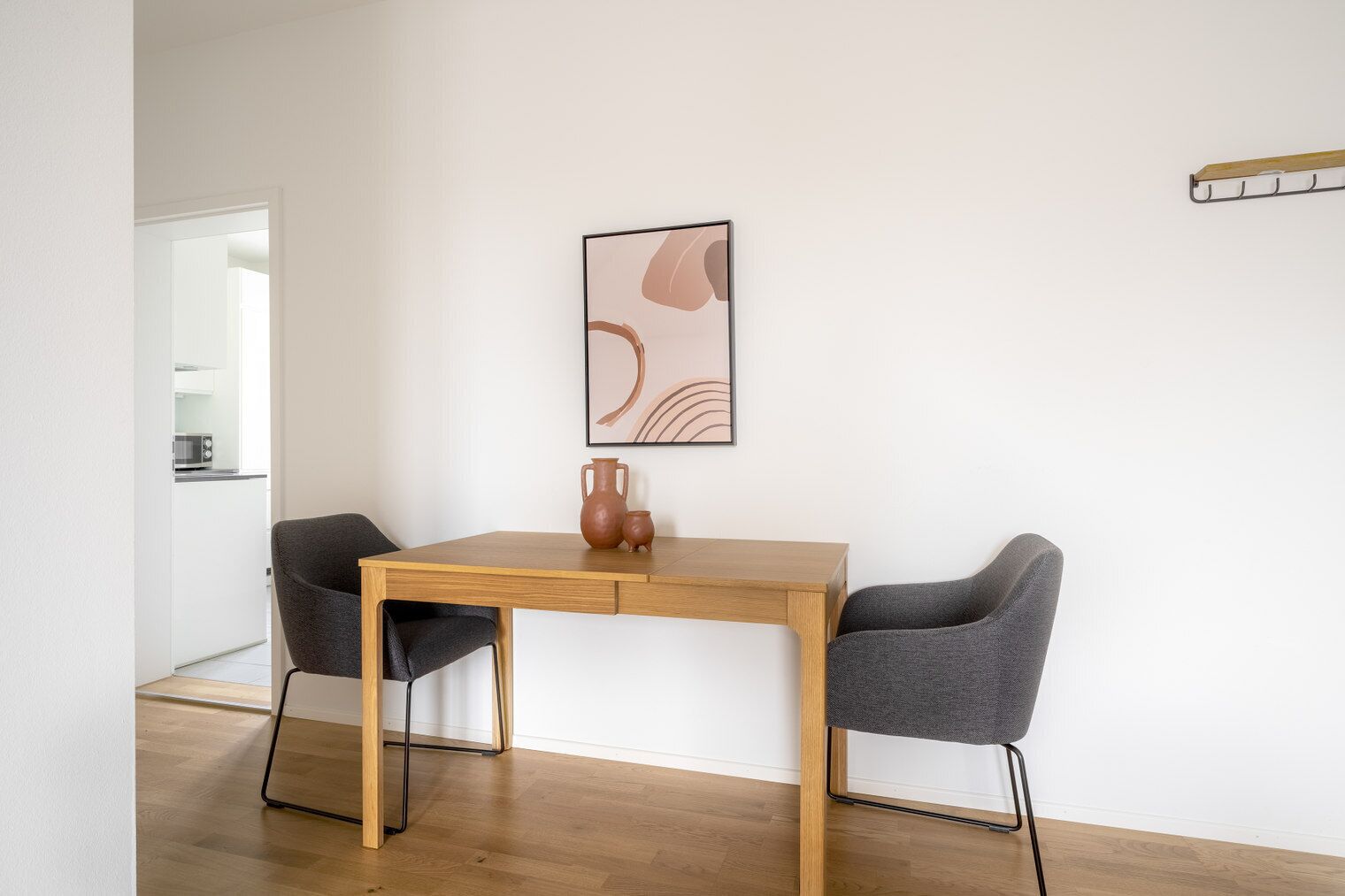 Un espace repas moderne avec une table en bois et deux chaises grises, un vase et un tableau abstrait encadré accroché au mur.