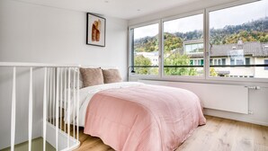 Ein Schlafzimmer mit großem Fenster, einem Bett mit rosa Decke, einer weißen Geländer und einem gerahmten Bild an der Wand.