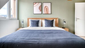 Un dormitorio moderno con cabecera de madera, una cama grande con colcha azul estampada, dos almohadas, dos obras de arte abstracto en la pared, lámparas de noche y una ventana con vistas a edificios.