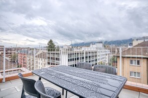Eine Dachterrasse mit Tisch und Stühlen, mit Blick auf eine Stadtlandschaft mit Gebäuden und Bergen im Hintergrund.